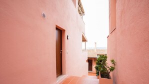 Un pasillo estrecho con paredes rosas y baldosas de terracota, que conduce a un edificio con tejado de tejas.