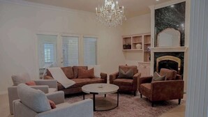 A living room with a fireplace, a chandelier, and a large window with blinds. - 4BR Sleep 12 | Family, Corp Stays | Palo Duro Cyn (Amarillo)