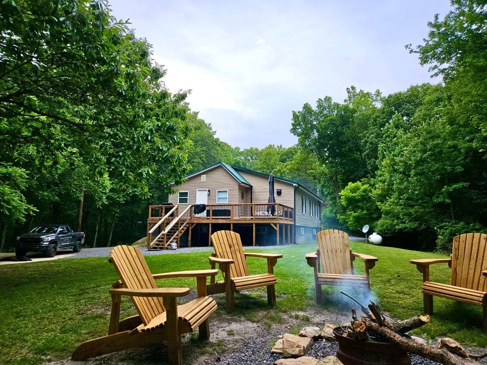 A cabin with a deck, surrounded by a grassy yard and trees. There are four wooden Adirondack chairs and a fire pit.
