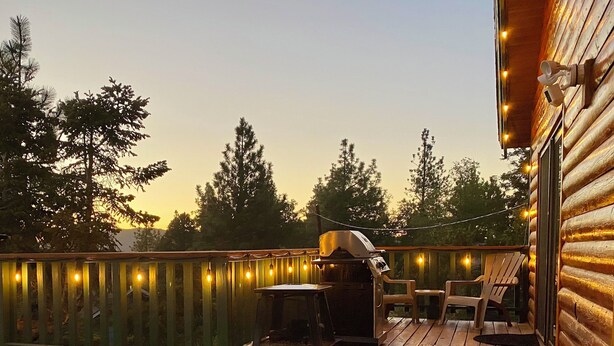 A wooden deck with a barbecue grill, outdoor seating, and string lights. - Wolf View Lodge (Big Bear City)