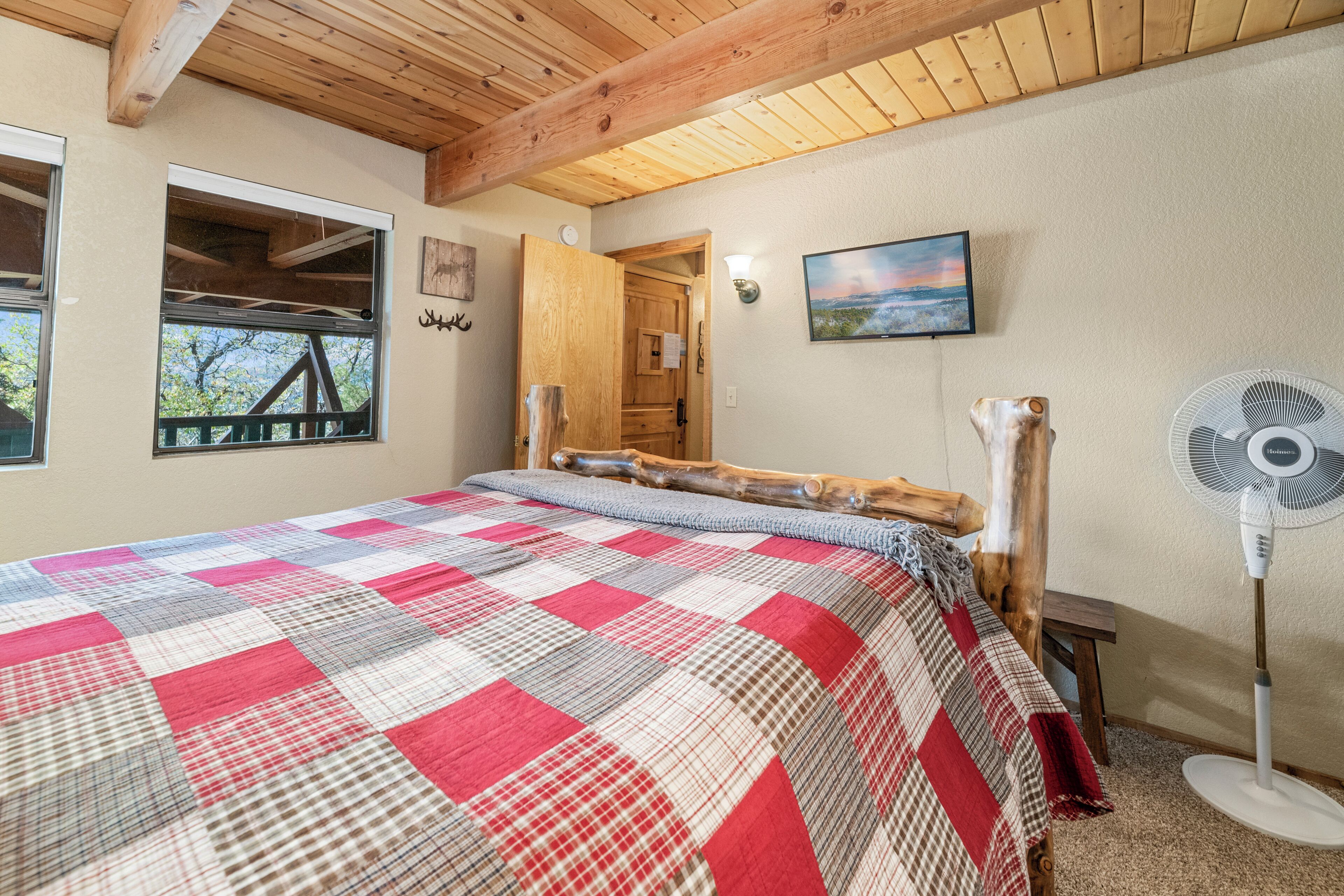 A bedroom with a wooden ceiling, a large bed with a red and white checkered bedspread, a wooden headboard, a standing fan, and a window with a view of trees.