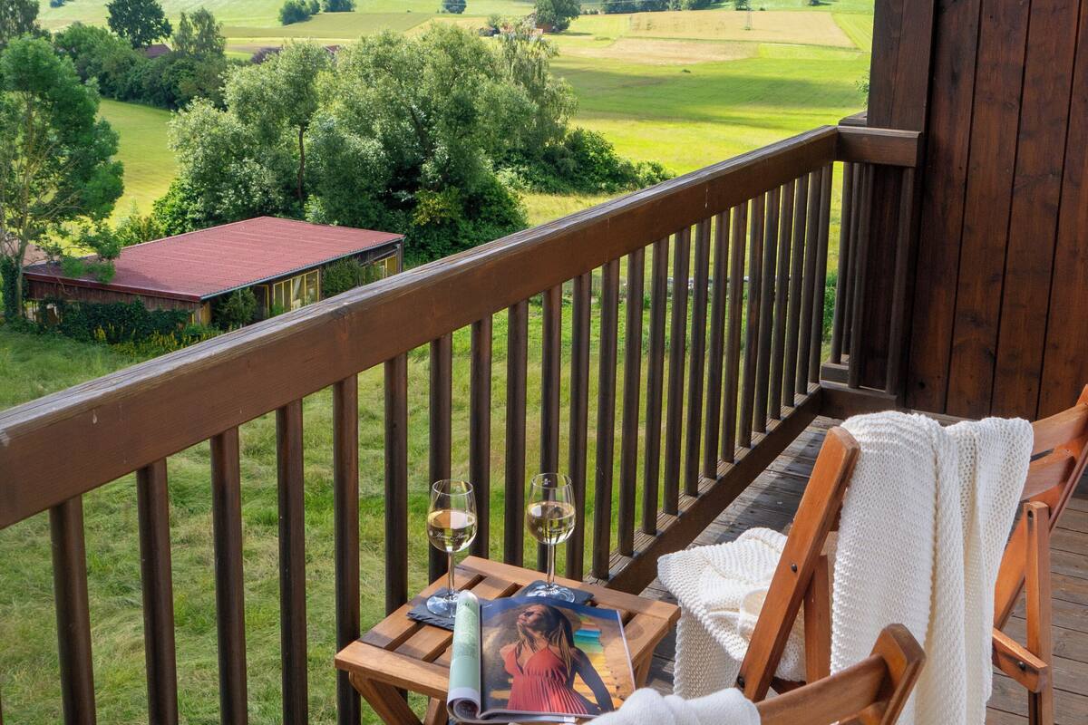 A wooden deck with a table and chairs, overlooking a green landscape with hills and a red-roofed building.