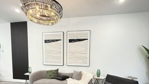 Een moderne woonkamer met een zwarte salontafel, een grijze bank, een zwarte bank en een beige bank. Twee ingelijste kunstwerken hangen aan de muur, een groene vaas op een bijzettafeltje, en een kroonluchter hangt aan het plafond.