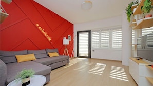 A modern living room with a red accent wall, a grey sofa with yellow cushions, a white coffee table with a plant, and a TV on a wooden stand.