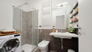 A modern bathroom with a glass shower enclosure, a bidet, a washing machine, and a sink with storage shelves.
