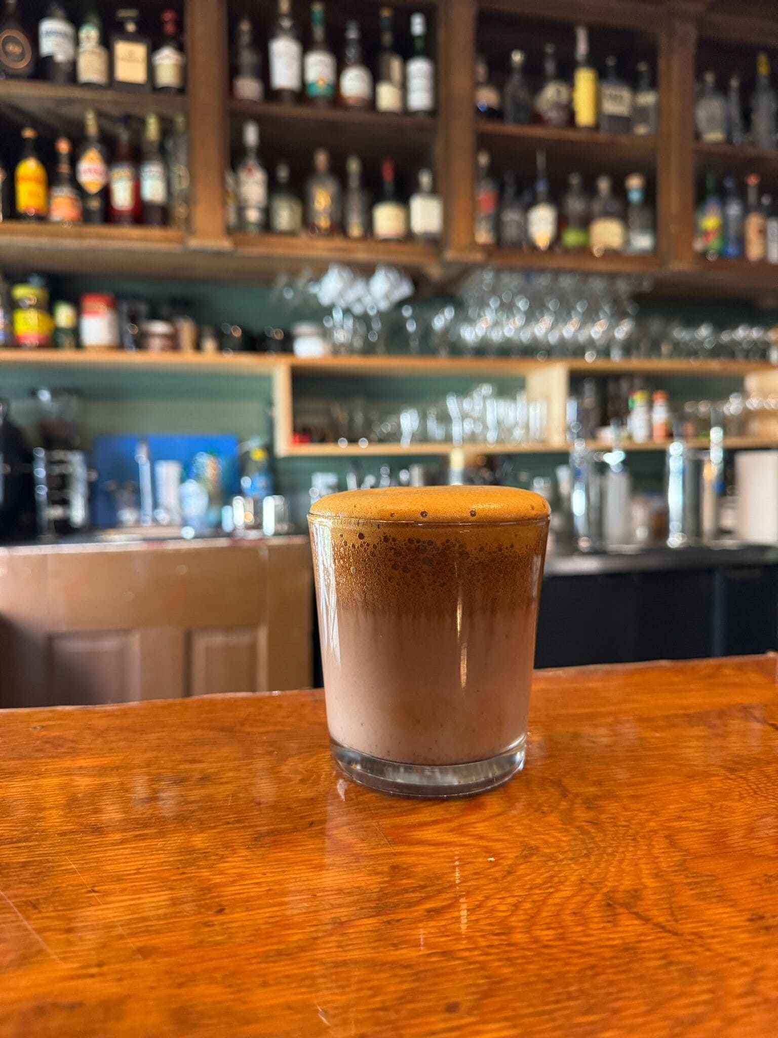A glass of a layered beverage on a wooden bar counter with shelves of bottles in the background.