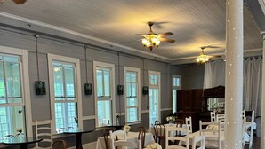 Una habitación amplia con suelos de madera, mesas y sillas blancas, ventanas grandes e iluminación decorativa.