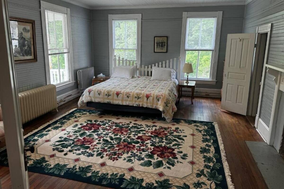 A bedroom with a large floral rug, a bed with a white headboard, two windows with white shutters, and a radiator.