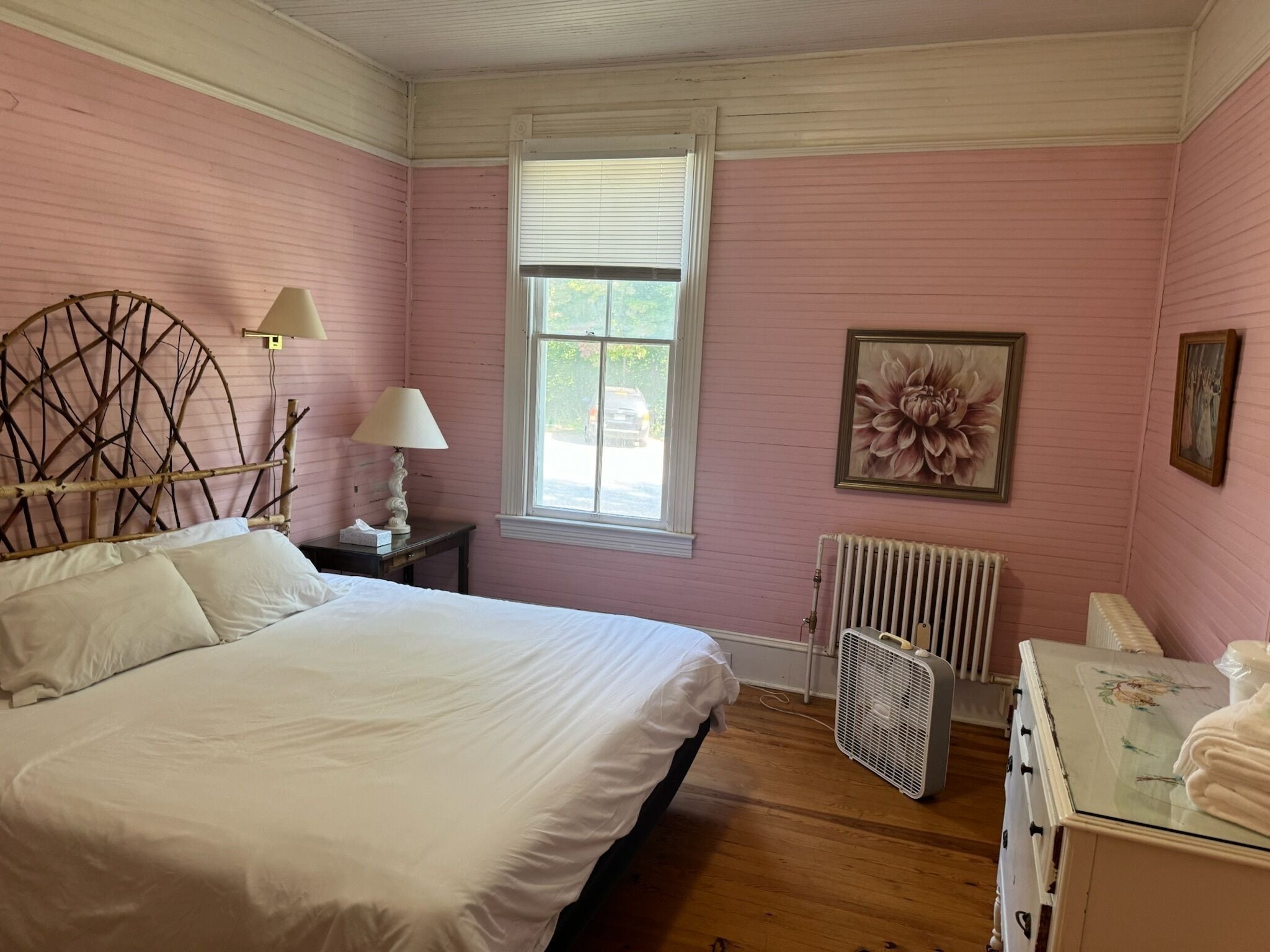 A bedroom with a bed, a radiator, a window, and a wall-mounted fan.