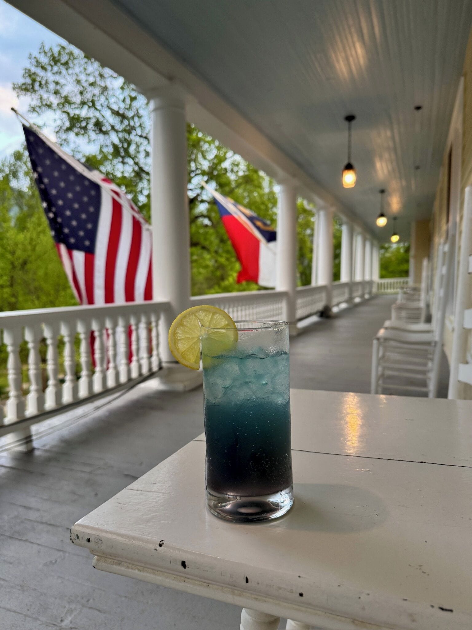 A glass of blue beverage with a lemon slice on a white table on a covered porch with flags in the background.