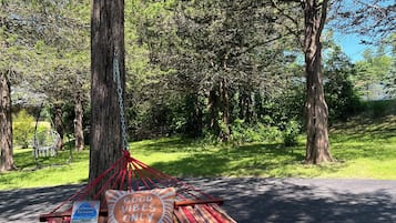 Un hamac est attaché à un arbre dans un parc, avec un livre intitulé « Good Vibes Only » et une pancarte indiquant « maison d’été ».