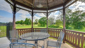 A gazebo with a round metal table and four chairs, surrounded by a wooden fence with decorative cutouts, overlooking a green landscape.