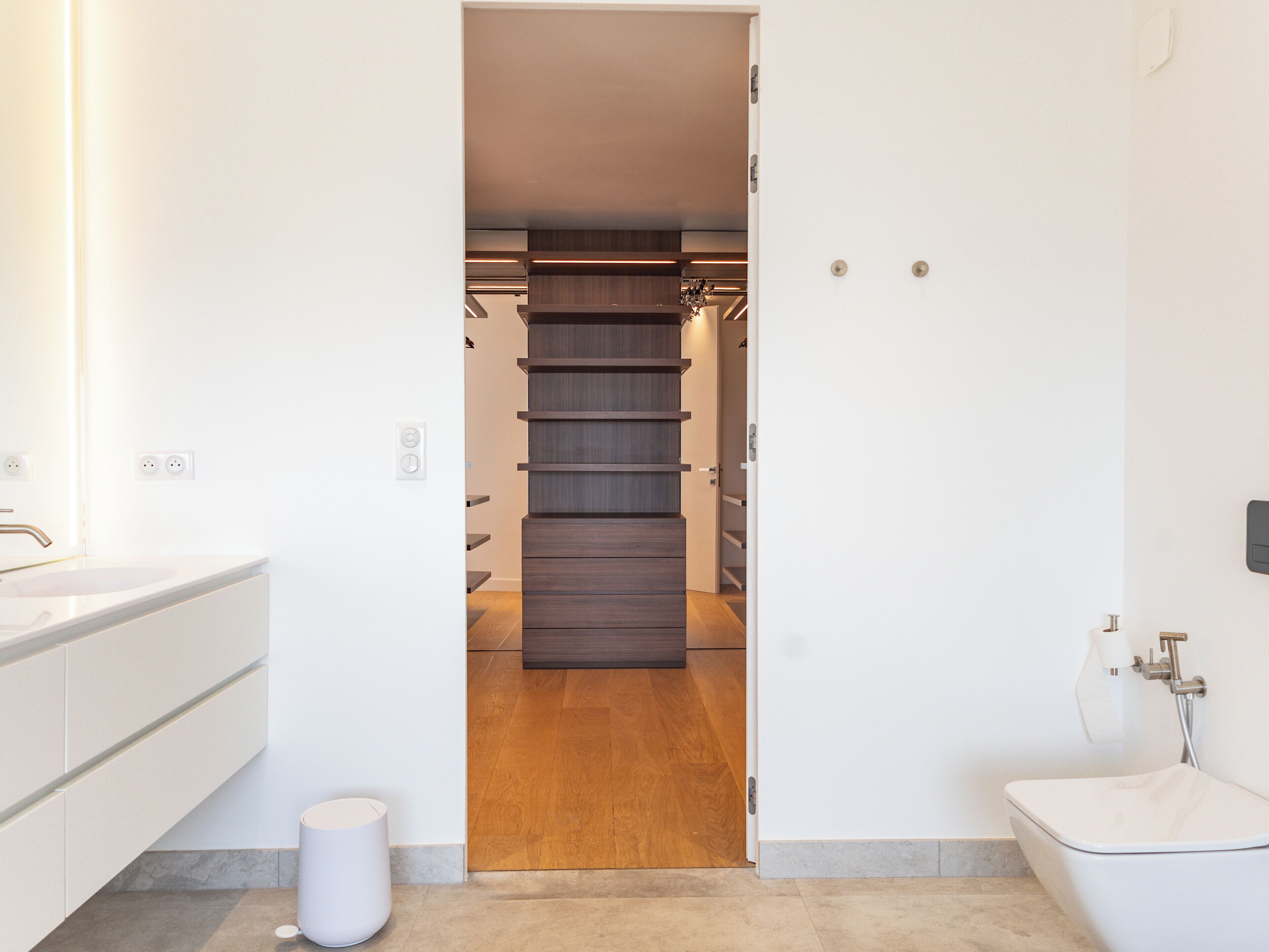 A modern bathroom with a white sink, a toilet, and a shower. There is a wooden floor and a white trash can.