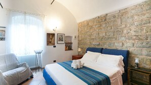 Un dormitorio con pared de piedra, una cama con cabecera azul, una silla, una mesita y un radiador.