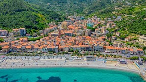 Vista aérea de un pueblo costero con playa, edificios de techos rojos y una ladera verde y densa en el fondo.