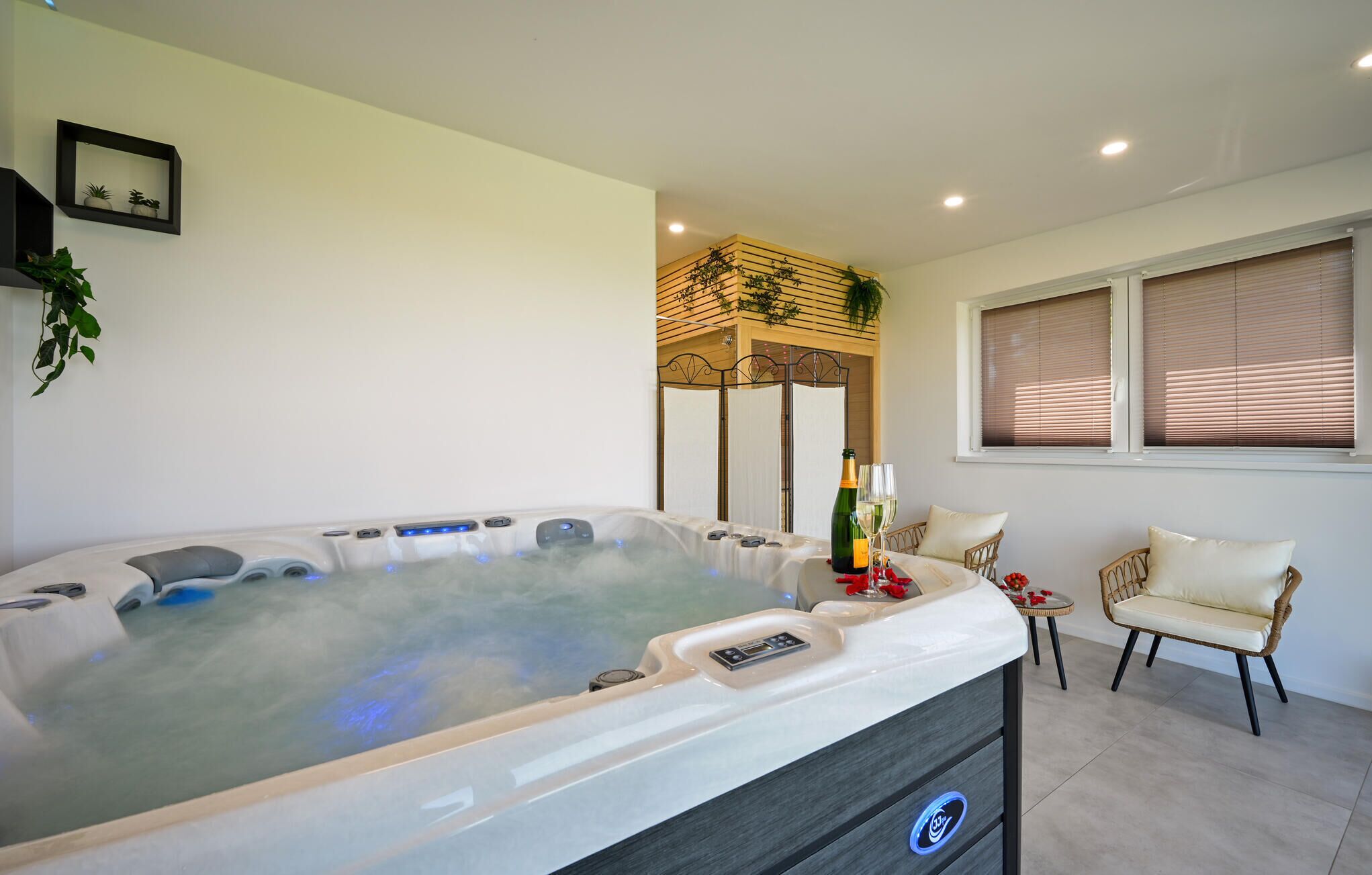 A modern bathroom with a large hot tub, a small table with a bottle and glasses, and two wicker chairs.
