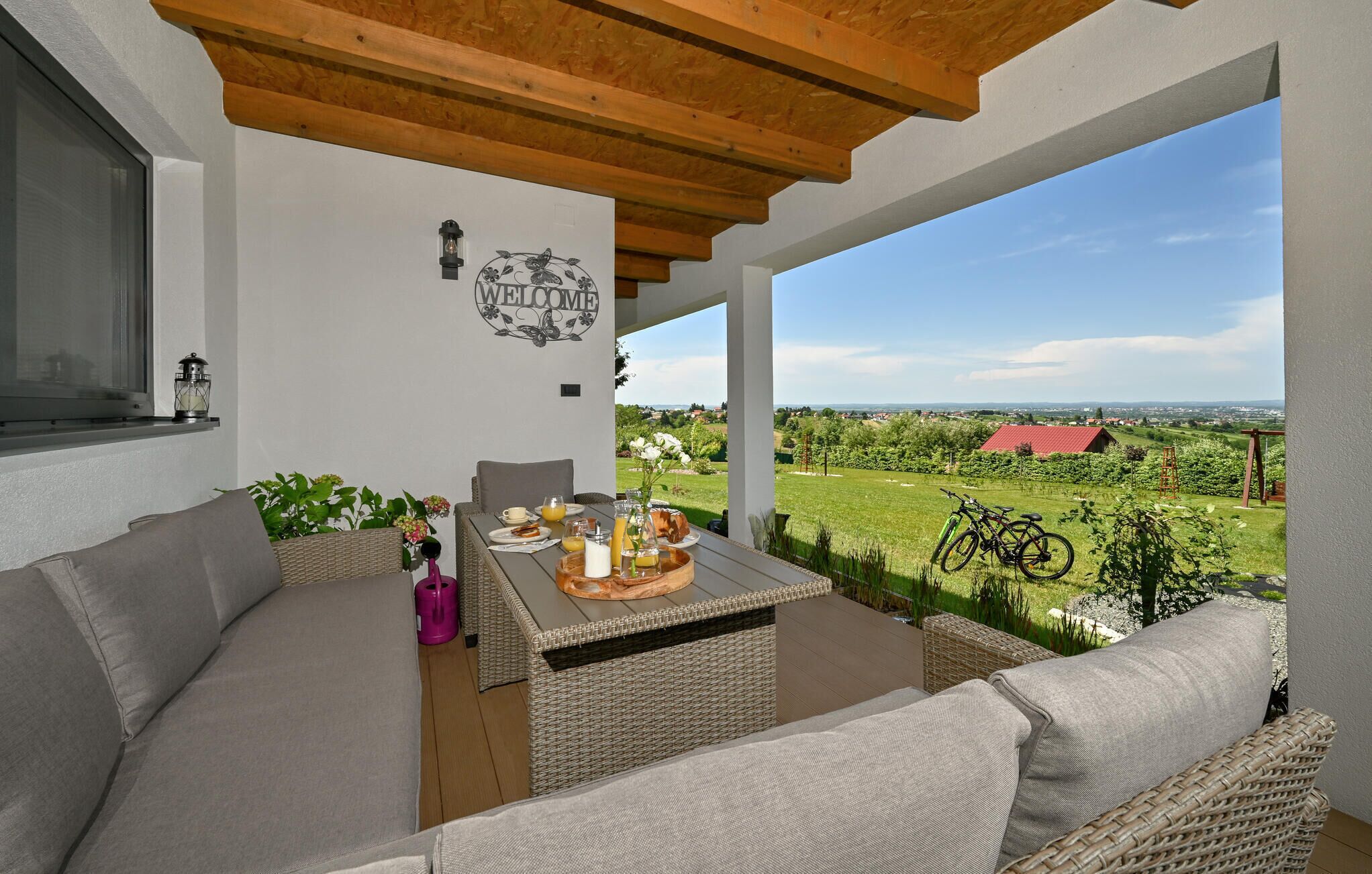 A covered outdoor seating area with wicker furniture, a table set with breakfast items, and a view of a rural landscape.