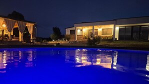 Een zwembad met een blauwe gloed, een overdekt terras met stoelen, en een modern gebouw met grote ramen en een stenen gevel.