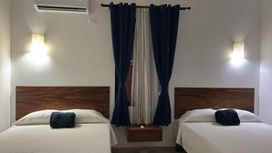 Ein Hotelzimmer mit zwei Betten, jeweils mit einem Holz-Kopfteil, weißer Bettwäsche und einem dunkelblauen Kissen. Es gibt ein Fenster mit blauen Vorhängen und einen kleinen Nachttisch zwischen den Betten.