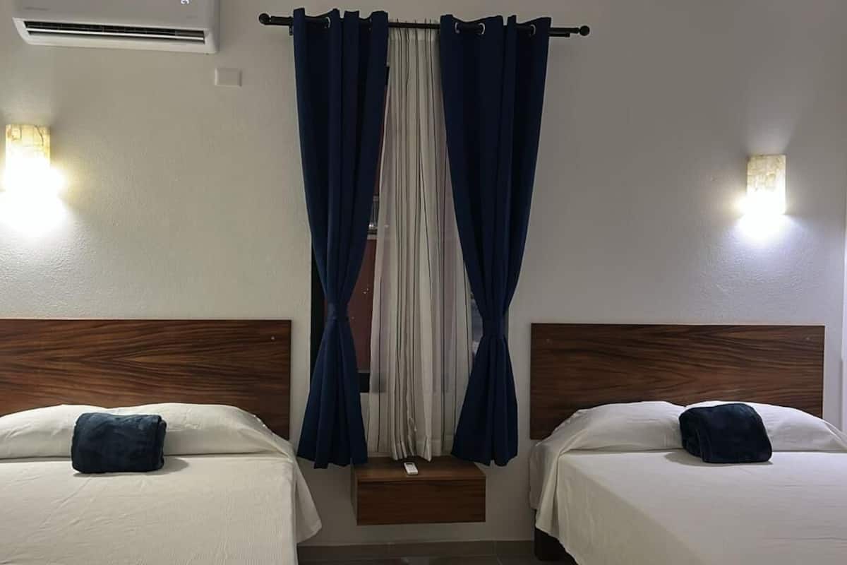 A hotel room with two beds, each with a wooden headboard, white bedding, and a dark blue pillow. There is a window with blue curtains and a small wooden nightstand between the beds.