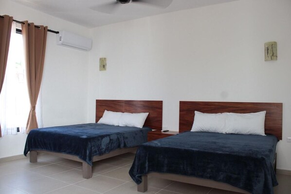 A room with two beds, each with a wooden headboard and a dark blue bedspread. There is a window with beige curtains, and an air conditioning unit mounted on the wall. - Hotel COCO (San Blas)