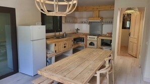 Keittiössä on puukehikoiset kaapit, valkoinen jääkaappi, puinen ruokapöytä ja katosta roikkuva valaisin.
