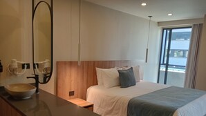 Una habitación de hotel moderna con una cama grande, cabecera de madera, mesita de noche, un espejo amplio y vista al mar a través de un ventanal.