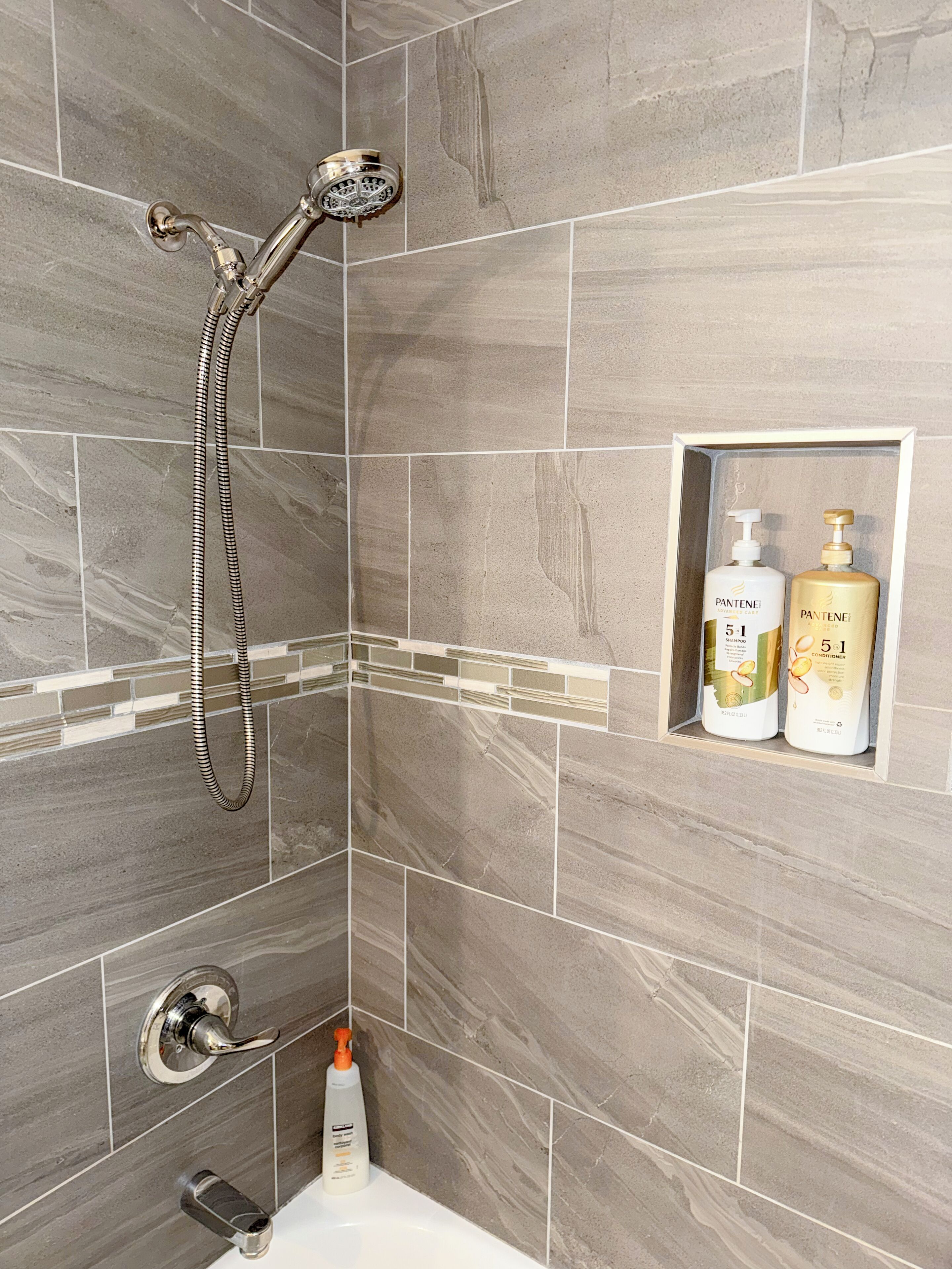 Ducha con cabezal de ducha manual, grifo empotrado en la pared y botella de champú y acondicionador sobre una repisa.