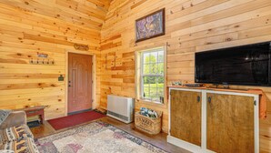 Interior acogedor de una cabaña con paredes de madera, un televisor, chimenea y un ventanal grande.