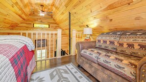 Acogedora habitación de cabaña de madera con techo de madera, cama con colcha roja y blanca, sofá estampado y una mesita con lámpara.