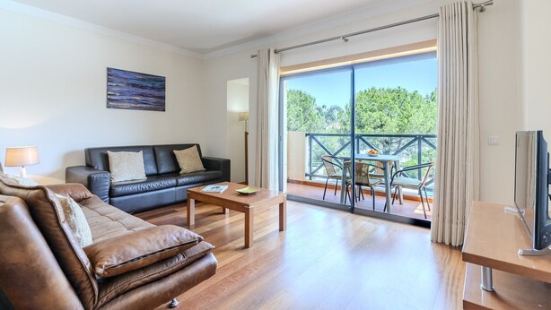 A modern living room with a sofa, a coffee table, and a TV stand. There is a balcony with a table and chairs.