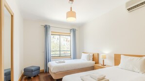 A hotel room with two beds, a small seating area, a window with curtains, a bedside table with a lamp, and an air conditioning unit.