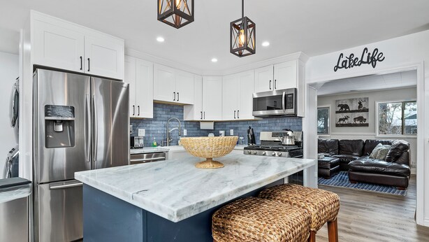 A modern kitchen with white cabinets, a stainless steel refrigerator, a marble island, and a woven stool. - Boulder Bay Bungalow (Big Bear Lake)