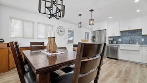 A modern kitchen with white cabinets, a stainless steel refrigerator, and a dining area with a wooden table and chairs. - Boulder Bay Bungalow (Big Bear Lake)