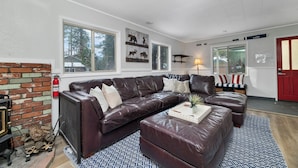 A modern living room with a sectional sofa, a fireplace, and a large window overlooking a forested area. - Boulder Bay Bungalow (Big Bear Lake)