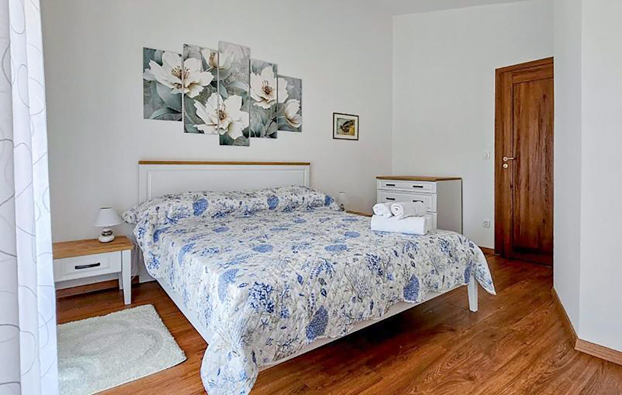 Ein Schlafzimmer mit einem Bett, einem Nachttisch, einer Kommode, einer Tür und einer Wand mit einem Blumenbild.