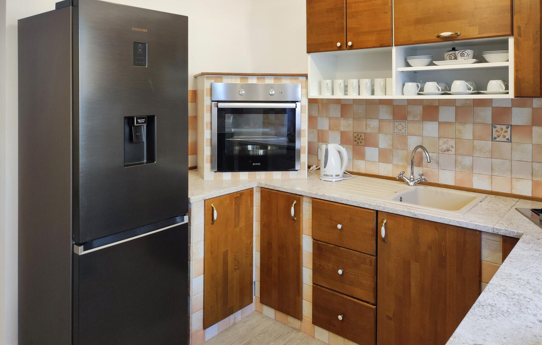 A compact kitchen with a stainless steel refrigerator, a built-in oven, wooden cabinets, and a tiled backsplash.