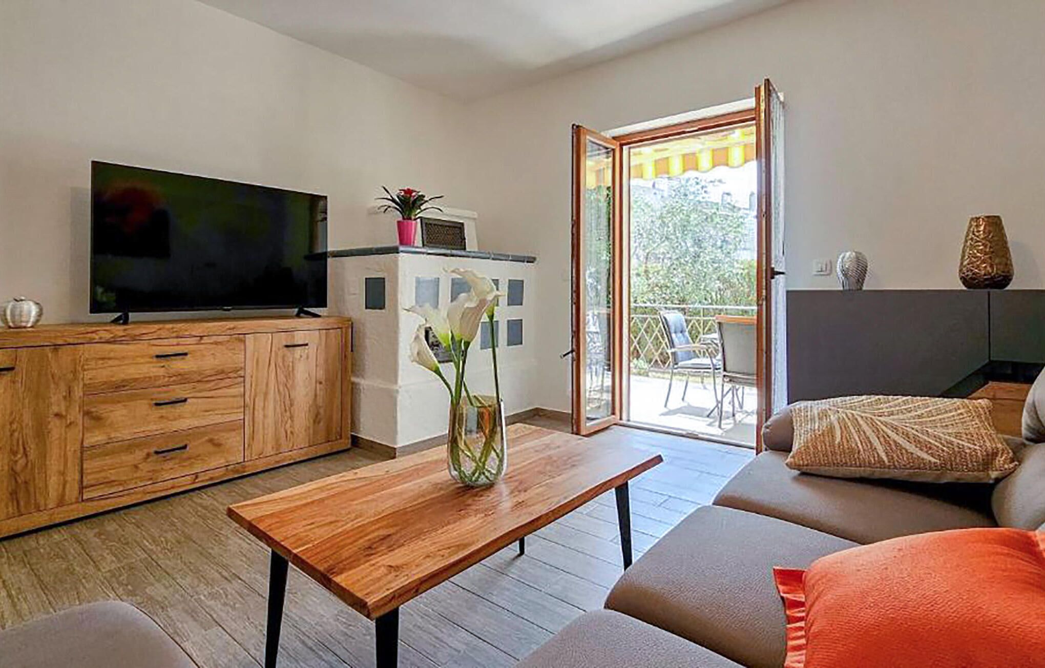 A modern living room with a wooden cabinet, a flat-screen TV, a coffee table with a vase of flowers, a sofa with cushions, and a sliding glass door leading to an outdoor area.