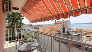 Ein Balkon mit Glastisch und Stühlen, einer roten Markise und Blick auf das Meer und Gebäude.