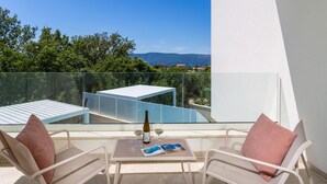Een balkon met twee stoelen, een tafel en een fles wijn, met uitzicht op een modern gebouw en groen.