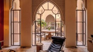 A room with a view of an outdoor patio, featuring a round table and chairs, a black leather chair, and ornate arches. - Riad MAYAN : Heated Pool & GYM (Marrakesh)