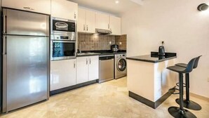A modern kitchen with stainless steel appliances, a built-in oven, a microwave, and a washing machine. There is a small bar area with two stools.