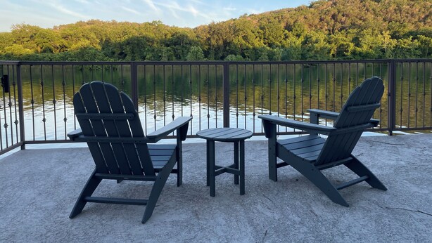 Eine Terrasse mit zwei Adirondack-Stühlen und einem kleinen Tisch, mit Blick auf einen ruhigen See und einen bewaldeten Hügel im Hintergrund.