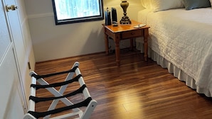 Ein Schlafzimmer mit Holzboden, einem Bett mit weißer Bettwäsche, einem Nachttisch mit Lampe, einem Fenster mit Jalousien und einem weißen Stuhl.