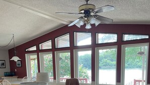 Ein geräumiges Wohnzimmer mit Deckenventilator, großen Fenstern, einem braunen Ledersessel, einem blauen Hocker und einem Essbereich mit Tisch und Stühlen.