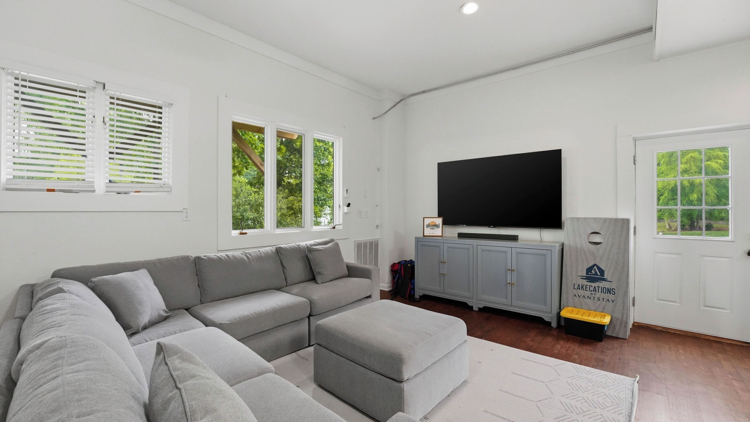 A modern living room with a sectional sofa, a flat-screen TV, grey cabinets, and large windows with blinds.