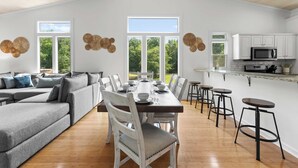 A spacious dining area with a long table, white chairs, and a bar area with stools. - Cove's End by Avantstay Dock, Game Room, Fire Pit & Forest Views (Mooresville)