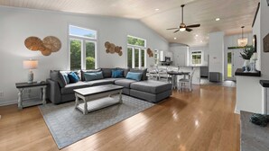 A spacious living room with a gray sectional sofa, a coffee table, and a dining area with a kitchen in the background. - Cove's End by Avantstay Dock, Game Room, Fire Pit & Forest Views (Mooresville)