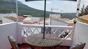 A balcony with a round wooden table and two chairs, a white railing, and a view of buildings and hills in the background.