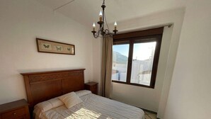 A bedroom with a wooden headboard, a large bed with a quilt, a nightstand, a bedside table, a chandelier, and a window with a view of buildings and mountains.